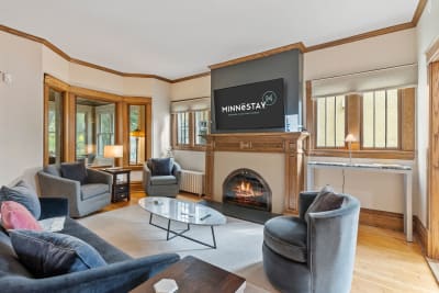 A stylish living room with a fireplace, sofa, armchairs, and a television displaying the Minnestay logo.