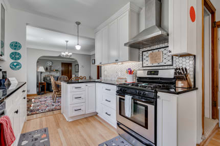 A modern kitchen with white cabinetry and a dark countertop, featuring a stove and dining area in the background.