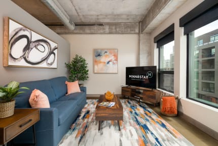 A stylish living room featuring a blue sofa, colorful artwork, and a TV displaying Minnestay logo.