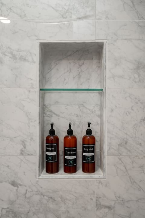 Shower niche featuring amber bottles of shampoo, conditioner, and body wash against a white marble backdrop.