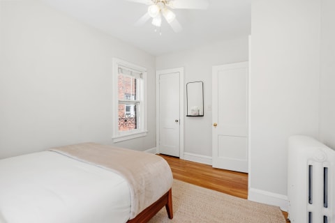A minimalist bedroom featuring a white bed with a beige throw, wooden floor, and natural light from a window.