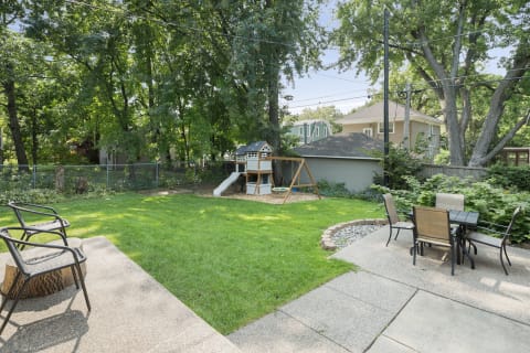 A spacious backyard with a playset, swing, and outdoor dining area surrounded by trees.