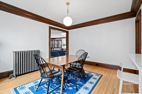 A dining room featuring a wooden table, black chairs, and a blue patterned rug, with a vintage radiator and pendant lighting.