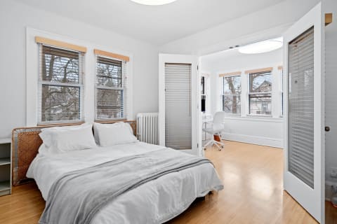 A cozy bedroom with a rattan bed, white bedding, and a desk area with windows.