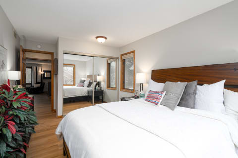 A well-decorated bedroom featuring a bed with white bedding and colorful pillows, side tables, and large windows.
