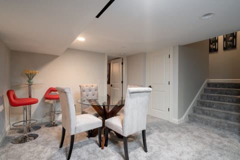 A modern basement space with a round glass dining table and plush chairs, accentuated by red bar stools and a staircase in the background.
