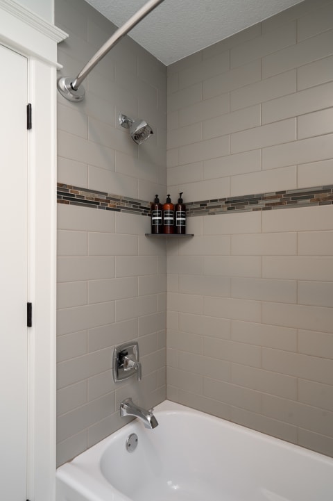 A modern shower area featuring gray tiles, a chrome showerhead, and amber bottles on a shelf.