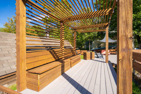 An outdoor wooden patio featuring a slatted pergola, benches, and green plants under a blue sky.