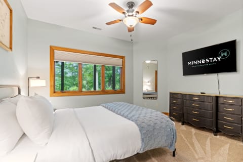 A contemporary bedroom featuring a bed with white bedding, a blue blanket, a dark dresser, a TV, and a view of trees outside.