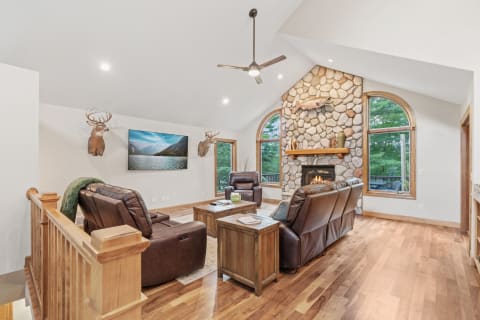 A cozy living room featuring a stone fireplace, leather chairs, and large windows with views of nature.