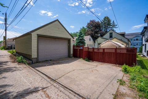 An alley featuring a light green garage, a red wooden fence, and surrounding houses under a blue sky.