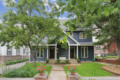 A two-story dark blue house with white trim, surrounded by greenery and pink flowers in pots.
