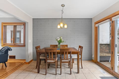 A dining room featuring a wooden table and chairs, with a vase of flowers and natural light from a sliding door.