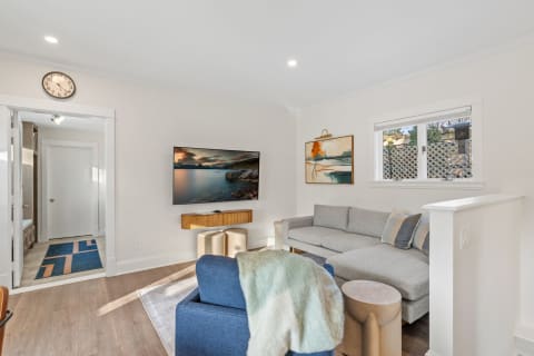A cozy living room featuring a wall-mounted TV, light gray sofa, blue armchair with a throw, and artwork on the wall.