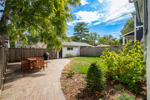A cozy backyard featuring a curved brick pathway, wooden dining set, grill, and greenery under a bright sky.