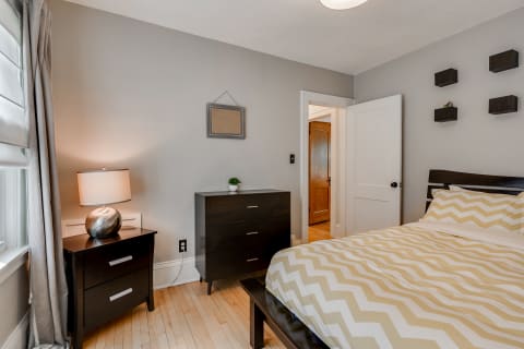 A modern bedroom with gray walls, a bed with zigzag patterned bedding, a nightstand with a lamp, and a dresser.