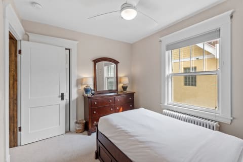 A cozy bedroom with a white bed, wooden dresser and mirror, and a window showing a view of an exterior wall.