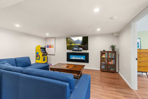 Interior view of a cozy basement living room featuring a blue sectional sofa, a television, an arcade game machine, and a decorative cabinet.