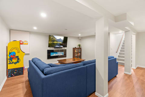 Modern basement with blue sectional sofa, arcade game, television, and electric fireplace.