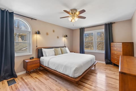 A modern bedroom featuring a white bed, wooden furniture, and large windows with dark curtains.