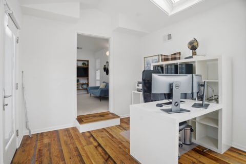 A contemporary home office with a white desk and wooden flooring, featuring dual monitors and natural light from a skylight.