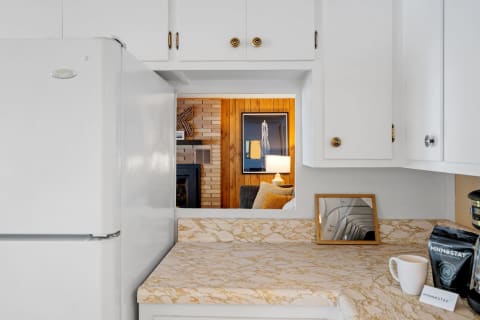 A kitchen with a white refrigerator, marble countertop, and a glimpse of a warm living room behind it.