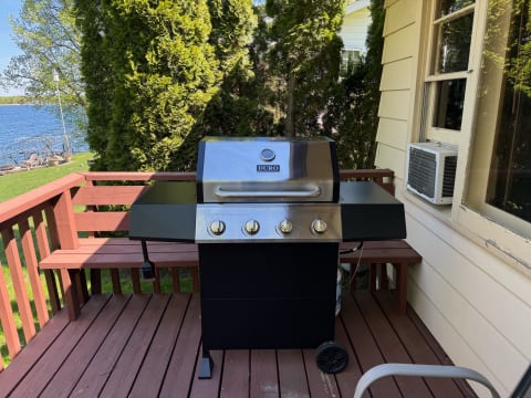 Gas grill on a wooden deck near a lake, surrounded by trees and a house.