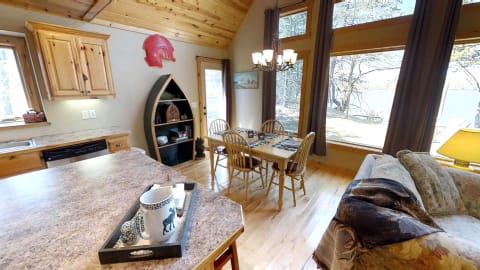 Interior view of a rustic kitchen and dining area with large windows overlooking trees and water.