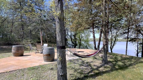 Secure hammock between trees near a lakeside patio with chairs and wooden barrels.