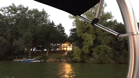 Evening view of a lit house from a boat on a calm lake, surrounded by trees.