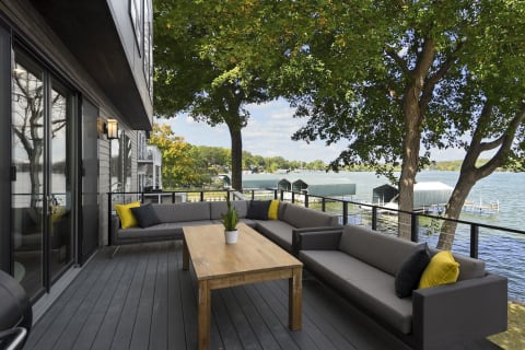 A stylish outdoor patio overlooking a lake, featuring gray sofas and a wooden table.