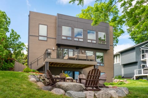 A contemporary house with large windows and a deck, surrounded by greenery and Adirondack chairs.