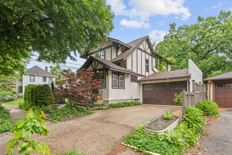 A two-story house with a pitched roof, surrounded by green trees and a gravel driveway.