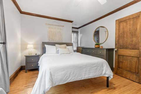 A bright bedroom with a neatly made bed, decorative pillows, a nightstand, and a dresser with a mirror.