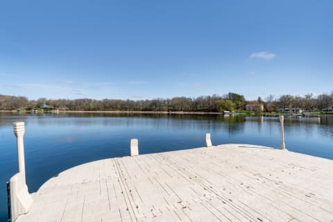 A serene view of a lake from a light-colored wooden dock with trees and houses in the background.