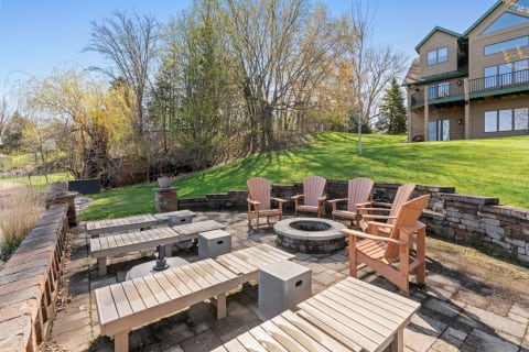 Outdoor patio area with wooden chairs and a fire pit, set against a backdrop of grass and trees.