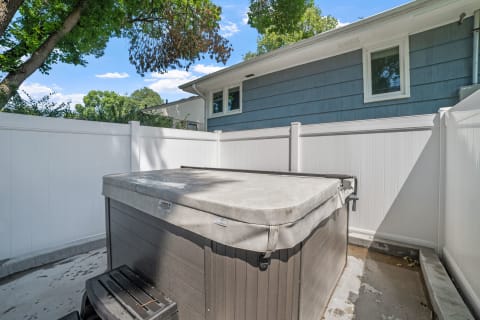 Hot tub surrounded by a white fence with a blue house and trees in the background.