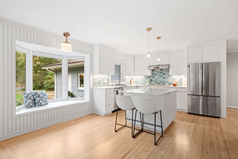 A bright and modern kitchen featuring white cabinetry, a bay window with seating, and a blue tiled backsplash.