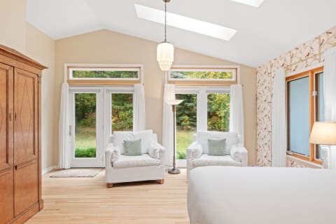 A cozy bedroom featuring white armchairs, a wooden wardrobe, and natural light coming through large windows.
