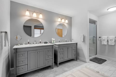 A modern bathroom with grey vanities, marble counter, and round mirrors.