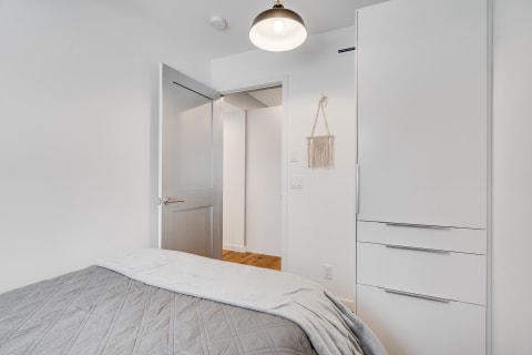 A minimalist bedroom featuring a bed with a gray blanket, an open door leading to a hallway, and a white cabinet.