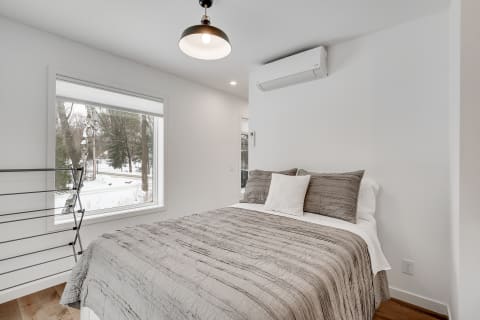 A modern bedroom with a double bed covered in a gray quilt, a drying rack, and large windows showing snow-covered trees.