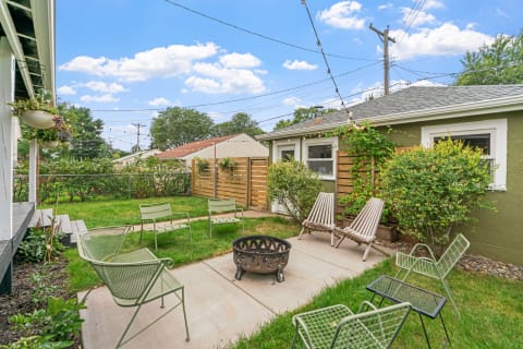 A peaceful backyard featuring a fire pit, chairs, and greenery under a blue sky.