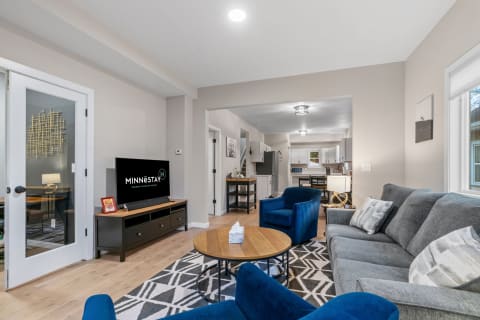 A cozy living room featuring a grey sofa, blue chairs, round coffee table, and an open kitchen in the background.