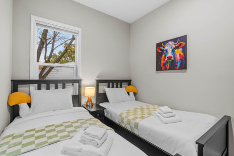 A cozy room with two single beds, a colorful cow painting, and a view of trees outside.