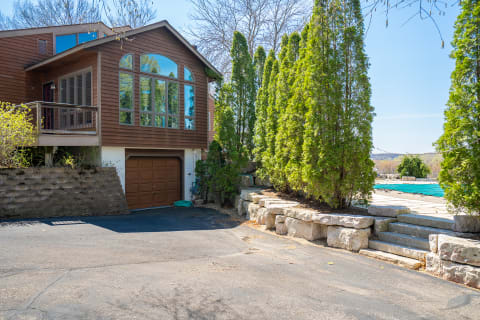 A brown wooden house with large windows and a paved driveway, surrounded by green trees and a pool area.