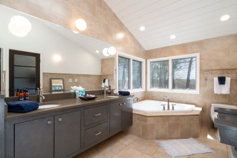 Spacious and modern bathroom featuring a dark gray vanity, a jetted bathtub, and large windows with a view of trees.