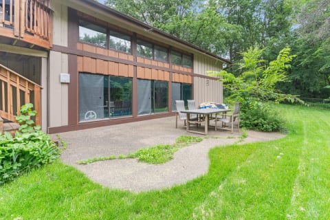 Patio area with a dining table and chairs next to a modern home set in a green landscape.