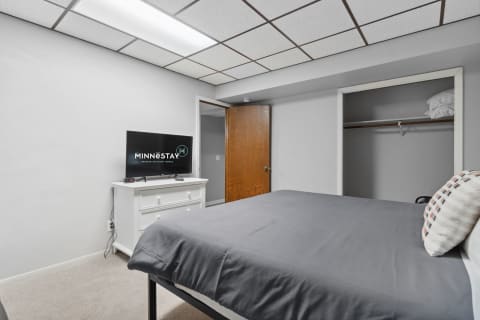 A neatly arranged bedroom with a gray bedspread, white dresser, and a TV displaying the MINNESTAY logo.