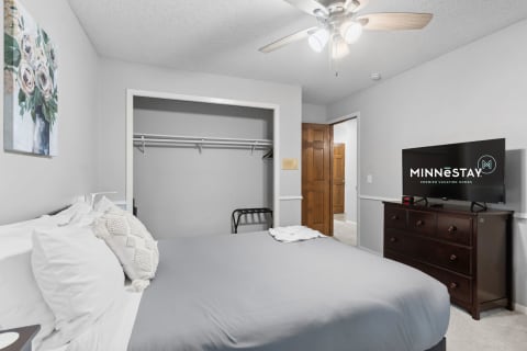 A bedroom featuring a gray color scheme, a bed with white pillows, a flower painting, a dresser with a TV, and an open closet.